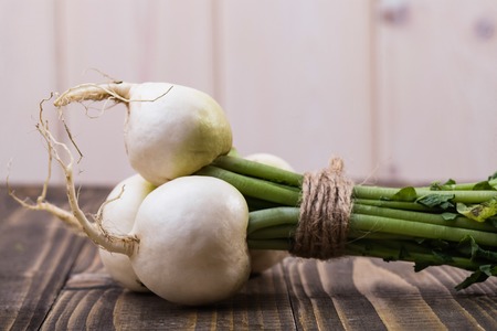 Ripe raw crude white round daikon with greens in bundle connected with twine on brown wooden table on white wall background, horizontal pictureの写真素材