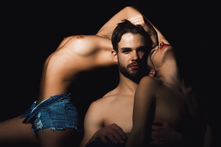 Three young sexy people of handsome bearded muscular macho man touching two sensual attractive girls lovers with beautiful body and naked chest close to each other indoor, horizontal pictureの写真素材