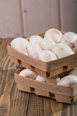 Two little wicker baskets with big fresh white champignons standing on each other on wooden table, vertical photoの写真素材