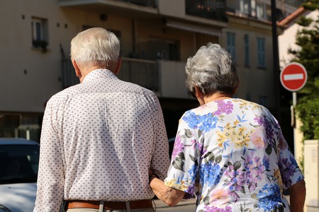 Photo back view of well-dressed silver-haired senior couple aged man and woman walking arm-in-arm in town on streetscape background, horizontal pictureの写真素材