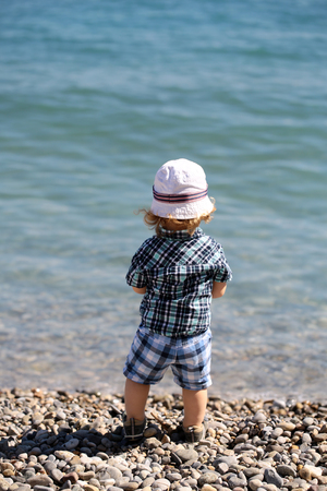Baby boy cute fair-haired blond kid tiny little child wearing checked shirt shorts and white hat standing on pebble beach on sunny day back view on beautiful seascape background, vertical pictureの写真素材