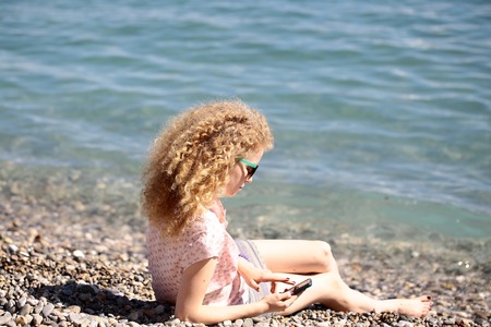 Woman one young with blond curly hair wearing sun glasses and playing with smart phone reclining on pebble beach on blue sea shore sunny summer day on blurred seascape background, horizontal pictureの写真素材