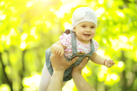 Baby girl smiling hazel-eyed kid tiny little child wearing white flower beanie hat lifted in air high by hands portrait outdoor on summer day on blurred sunny background, horizontal pictureの写真素材