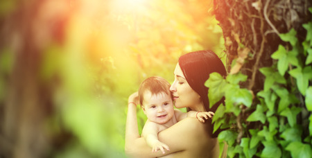 Baby and mother hugging kissing nude portrait sun spot outdoor summer nature on blurred green background, horizontal pictureの写真素材