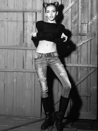 One straight slender funny young woman with cool hairstyle in torn jeans and black sweater with flexible body standing in studio on wooden backdrop black and white, vertical pictureの写真素材
