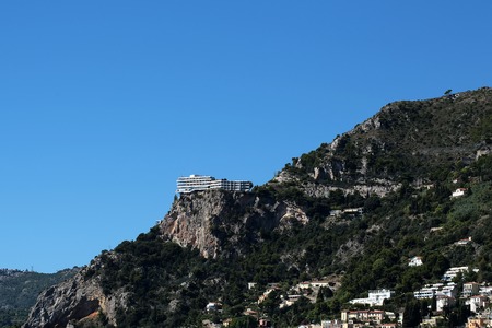 Monte Carlo, Monaco - September 20, 2015: mountain view cliffed coast rocky shore solitary white building hotel situated on edge of cliff on bright blue sky background, horizontal pictureのeditorial素材