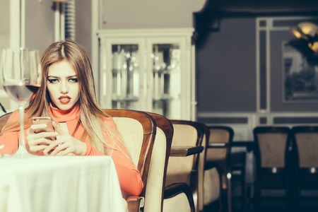 Elegant sexy young confident woman with long lush beautiful hair sitting in restaurant with mobile phone near wine glass in orange dress, horizontal pictureの写真素材