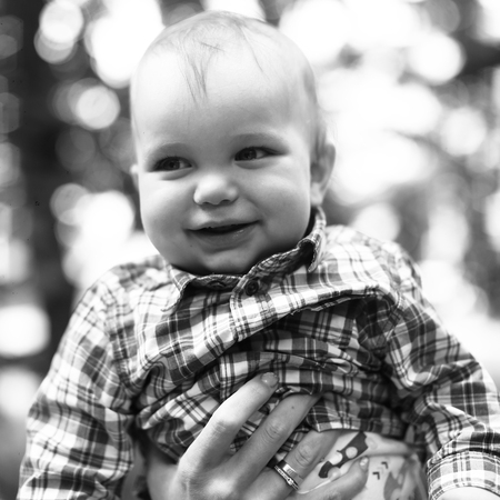 Portrait of smiling little boy in plaid shirt in mother's hands in air on blurred background, black...