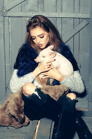 Beautiful young woman with stylish thin rim on hair sitting holding cute pink small pig pet on hands on wooden background, vertical pictureの写真素材