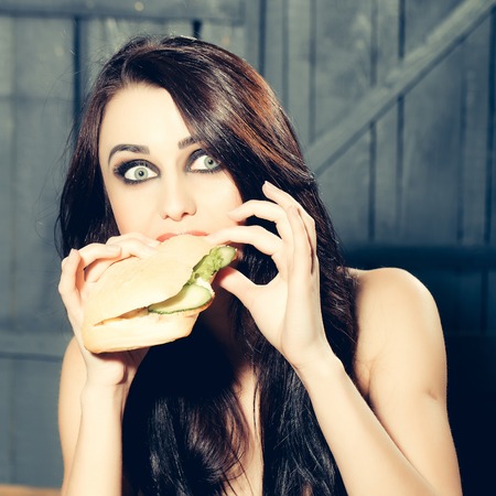 Emotional surprised young woman with long brunette hair and naked body eating big fresh tasty burger sitting indoor on wooden background, square pictureの写真素材