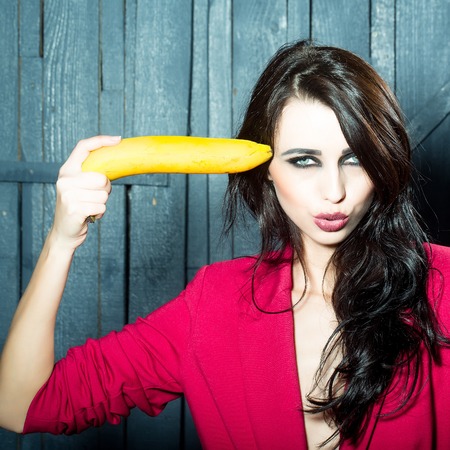 Sensual elegant emotional beautiful young woman with long lush brunette hair in stylish jacket holding banana as gun indoor on wooden background, square pictureの写真素材