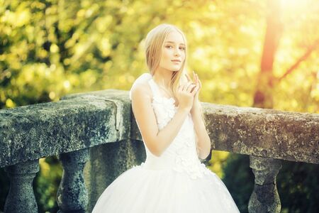 Attractive sensual thoughtful young bride girl with long blonde hair in white wedding dress standing outdoor near stone fence on natural background, horizontal pictureの写真素材
