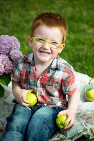 Happy laughing funny little red haired boy in stylish checkered shirt jeans and yellow glasses sitting outdoor on picnic with green apples near pink flowers on natural backdrop, vertical pictureの写真素材