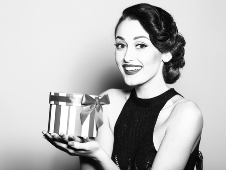Portrait of attractive sensual glamour retro elegant young adult smiling woman with classic hairstyle and red lips in evening dress holding present box indoor black and white, horizontal pictureの写真素材
