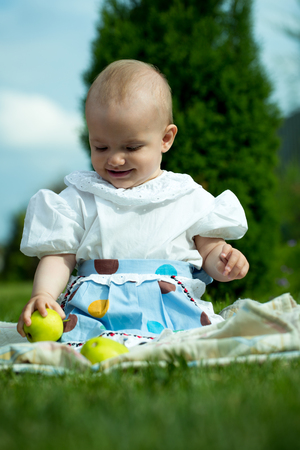 Happy smiling funny small baby girl in stylish dress sitting outdoor on picnic with green apples on natural grass backdrop, vertical pictureの写真素材