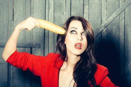 Sensual elegant emotional beautiful young woman with long lush brunette hair in stylish jacket holding banana as gun indoor on wooden background, horizontal pictureの写真素材