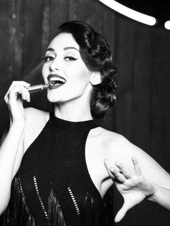Attractive sensual fashionable retro elegant young adult smiling woman with classic hairstyle and red lips holding lipstick in evening dress indoor on wooden background, vertical pictureの写真素材