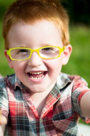 Portrait of funny happy smiling cute little boy with red hair in checkered shirt and yellow glasses sunny day outdoor on blurred natural background, vertical pictureの写真素材
