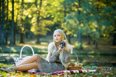 Stylish sensual thoughtful beautiful young blond girl with long hair in fashionable spring dress sitting on picnic plaid with basket of fruits in park outdoor on natural background, horizontal pictureの写真素材