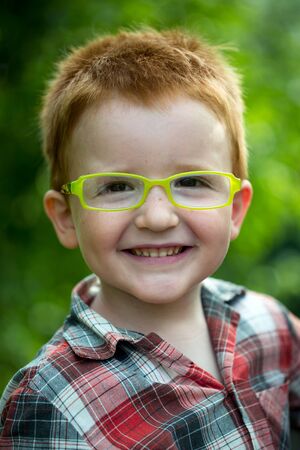 Portrait of funny happy smiling cute little boy with red hair in checkered shirt and yellow glasses sunny day outdoor on blurred natural background, vertical pictureの写真素材