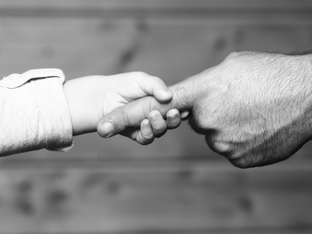 Closeup of two touching hands of small baby boy holding finger of male father as symbol of family love and trust on blurred wooden background, horizontal pictureの写真素材