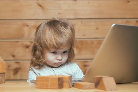 Cute funny little baby boy with long blonde curly hair playing on computer and mobile phone near toy building blocks indoor on wooden background, horizontal pictureの写真素材