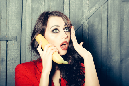 Sensual elegant emotional beautiful young woman with long lush brunette hair in stylish jacket holding banana as phone indoor on wooden background, horizontal pictureの写真素材