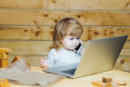 Cute funny little baby boy with long blonde curly hair playing on computer and mobile phone near toy building blocks indoor on wooden background, horizontal pictureの写真素材