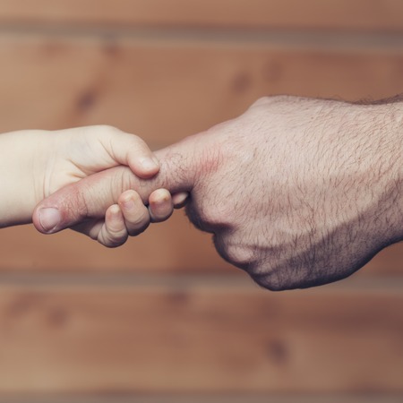 Closeup of two touching hands of small baby boy holding finger of male father as symbol of family love and trust on blurred wooden background, square pictureの写真素材