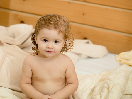 Cute happy beautiful smiling playful child boy with wet hair sitting in hothouse bath white fluffy towel naked indoor on wooden background, horizontal pictureの写真素材