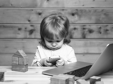 Cute funny little baby boy with long blonde curly hair playing on computer and mobile phone near toy building blocks indoor on wooden background, horizontal pictureの写真素材