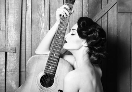 Sensual elegant retro glamour young adult woman with beautiful hair style red lips and bare shoulder holding acoustic string musical instrument of guitar indoor on wooden background, horizontal photoの写真素材