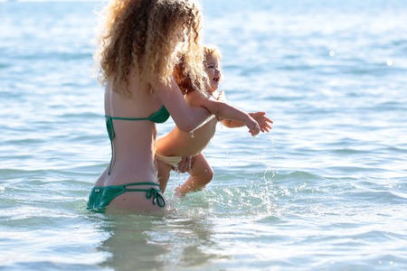 Young mother in bikini standing swimming and playing with male child boy in sea or ocean water sunny day outdoor on natural background, horizontal pictureの写真素材