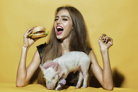 Beautiful young sensual woman and cute pink small pig pet in cloth eating tasty big fresh burger on...