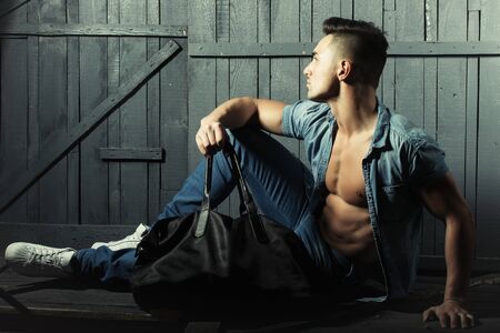 Handsome fashionable sexy sensual muscular young macho man with bare torso and stylish hair in jeans shirt holding sport bag indoor sitting on wooden background, horizontal pictureの写真素材