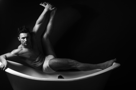Handsome sensual sexy young stylish man in white panties bare muscular torso and beautiful body indoor sitting on bathtub in bath room with raised leg on studio background, horizontal pictureの写真素材