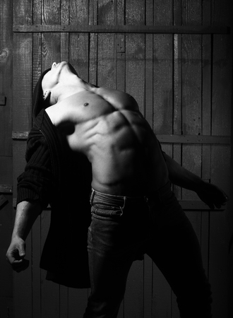 Sexual free stylish strong flexible muscular young macho man with bare torso in sweater standing indoor looking up on wooden background, vertical pictureの写真素材