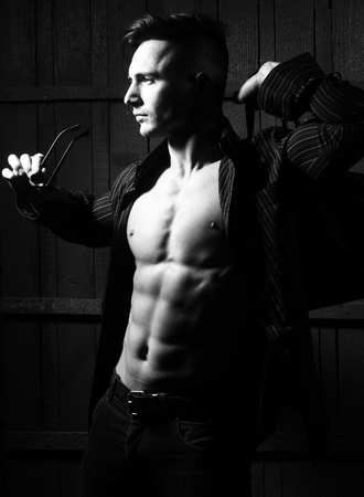 Sexy serious sensual muscular young macho man with bare torso and stylish hair in shirt sun glasses and bag standing indoor on wooden background, vertical pictureの写真素材