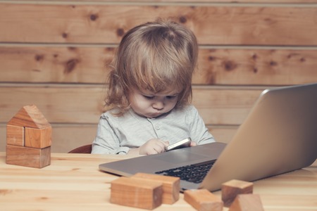 Cute funny little baby boy with long blonde curly hair playing on computer and mobile phone near toy building blocks indoor on wooden background, horizontal pictureの写真素材