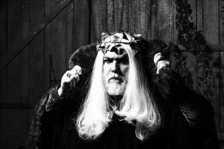 Bearded senior man in long white wig vine crown as Zeus god in fur coat indoor on wooden backgroundの写真素材
