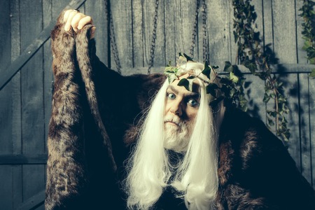 Bearded senior man in long white wig vine crown as Zeus god in fur coat indoor on wooden backgroundの写真素材