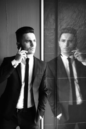 Man young handsome sensual elegant model in suit with skinny necktie open coat talks on mobile phone looks away hand in pocket reflects in mirror black and white on grey backgroundの写真素材