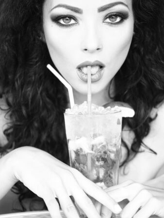 Closeup of beddable young curly brunette with bright makeup drinking cocktail with ice and lime from straw, black and whiteの写真素材