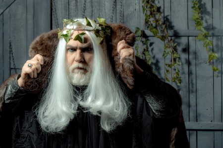 Bearded senior man in long white wig vine crown as Zeus god in fur coat indoor on wooden backgroundの写真素材