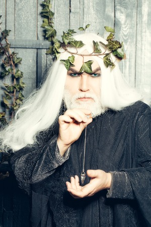 Bearded senior man wizard in long white wig vine crown as Zeus god with medallion on chain indoor on wooden backgroundの写真素材