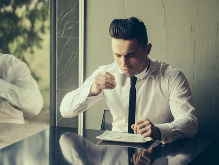 Man young handsome elegant model wears white shirt black skinny necktie sits at table drinks coffee reflects in glass window and looks in cup indoor on grey backgroundの写真素材