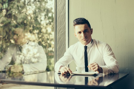 Man young handsome elegant model wears white shirt black skinny necktie sits at table drinks coffee reflects in glass window and looks in camera indoor on grey backgroundの写真素材