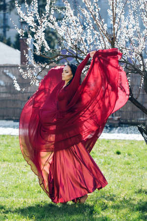 Young woman in beautiful red dress near apricot tree with blooming flowers.の写真素材