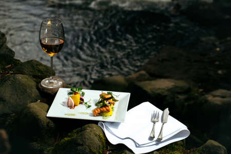Picnic near water, decorated fish meal with lemon, glass of wine, served foodの写真素材