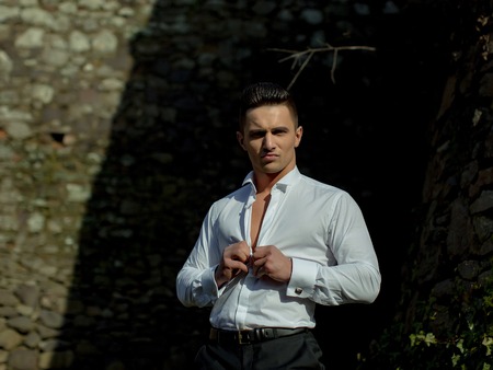 Man young handsome sensual model with plush lips undoes buttons on white shirt trousers looks at camera outside on masonry backgroundの写真素材